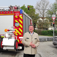 Verkehrsminister Christian Bernreiter steht auf einem Gehweg. Im Hintergrund ist ein Feuerwehrfahrzeug zu sehen, das an einer Ampel wartet.